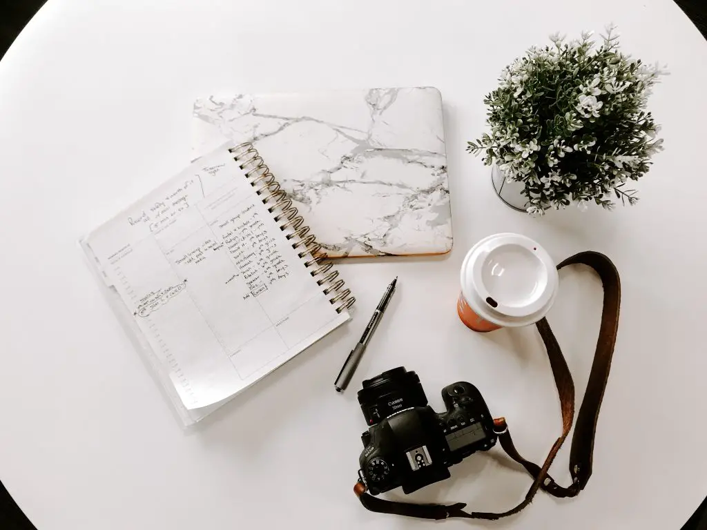 white notebook beside DSLR camera, coffee cup and plant