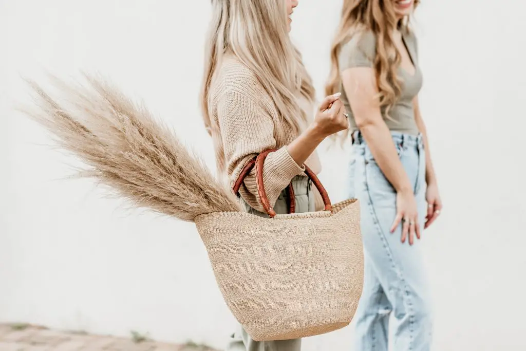 woman walking speaking to friend whilst holding brown wicker bag with pampass grass 