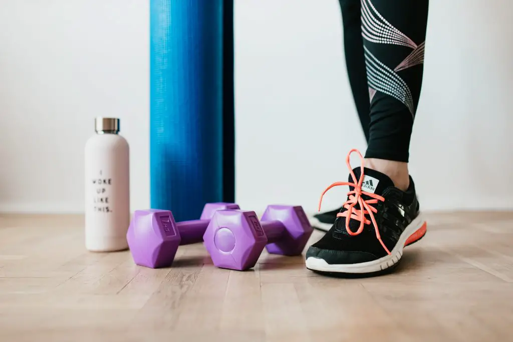 female in workout trainers standing beside water bottle, weights and yoga mat