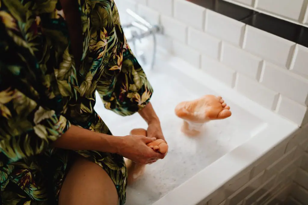 person sitting on side of bathtub massagin feet